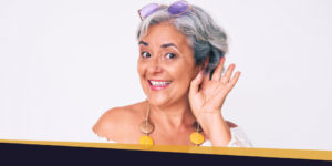 ApexBlog - Woman making a hearing gesture wearing a white off the shoulder shirt, a yellow and brown necklace with purple sunglasses on top of her head against a white background